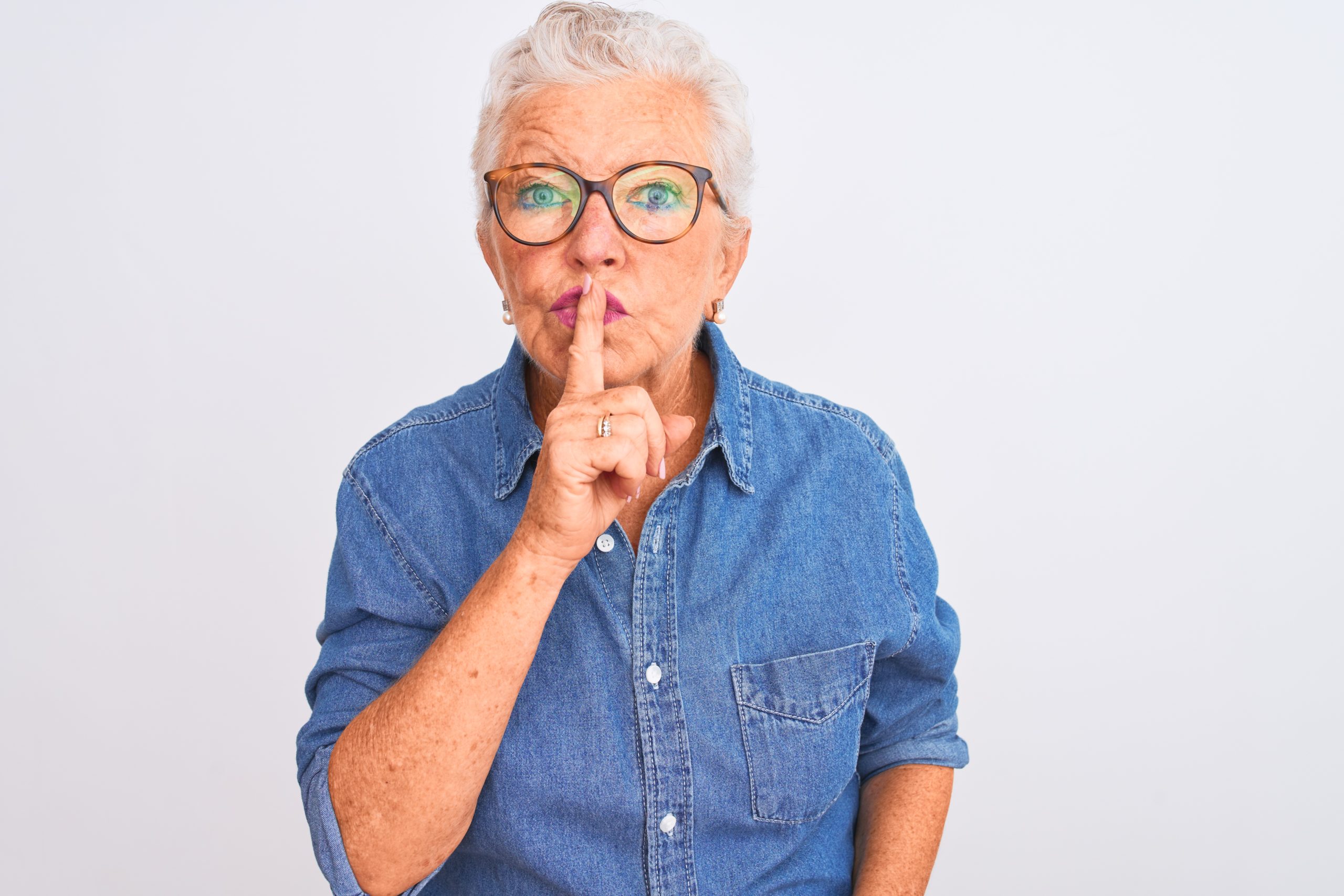 Senior grey-haired woman asking to be quiet with finger on lips. Pssst. Diskreter Immobilienverkauf.